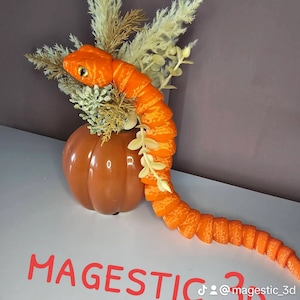 May include: A bright orange carrot snake sculpture sits on top of a brown ceramic pumpkin. The snake is made of many small carrot pieces, and the pumpkin is a decorative piece. The text "MAGESTIC 3D" is visible in the image.