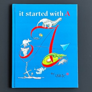 May include: A blue book cover with the title "it started with A" and a large red letter A with various animals and objects around it. The book is by Laura.