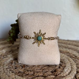 May include: A bracelet featuring a central gold-toned sunburst charm with a small round teal stone at its center. The charm is attached to a tan-colored woven cord, which includes small gold-toned beads on either side of the sunburst element.
