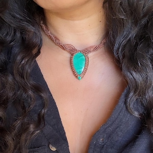 Peut inclure: Un collier composé d'une pierre de malachite verte en forme de goutte, sertie dans un cordon en macramé marron. Le cordon est finement tressé avec un motif complexe qui entoure le cou, maintenant la pierre centrale dans un cadre noué.