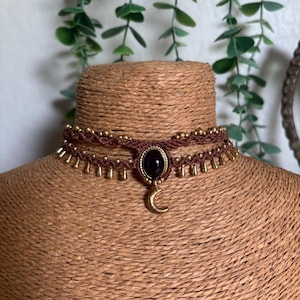 May include: A brown macrame choker necklace featuring a central oval dark purple gemstone in a gold-toned setting. The design includes small gold-toned beads along the top and bottom edges, with a gold-toned crescent moon charm hanging below.