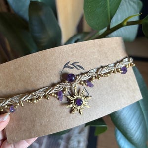 May include: A braided beige cord choker necklace featuring small purple amethyst beads and metallic gold-toned accents. A central gold-toned sun-shaped pendant is set with a round purple amethyst stone at its center, attached to the cord.