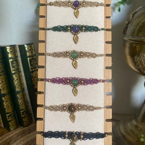 May include: Six macrame bracelets displayed on a vertical wooden stand. Each bracelet features a central gemstone, small metallic beads, and a dangling leaf-shaped charm. The cords vary in color, including beige, dark green, purple, and black.