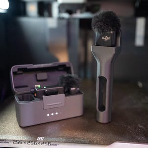 May include: A black microphone with a furry wind screen and a charging case. The microphone has the word "DJI" printed on it.