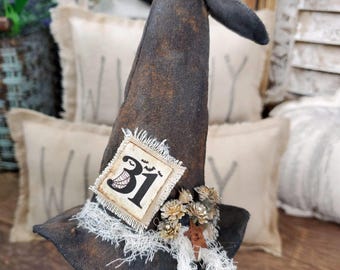 May include: A black witch hat with a frayed white trim and a brown fabric patch with the number "31" on it. The hat is decorated with brown and gold floral accents and is sitting on a wooden surface.