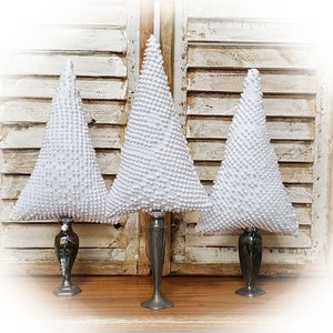 May include: Three white, cone-shaped decorative trees with textured surfaces. Each tree sits atop a silver-colored candlestick holder. The trees are arranged on a wooden surface in front of a distressed white shutter background.