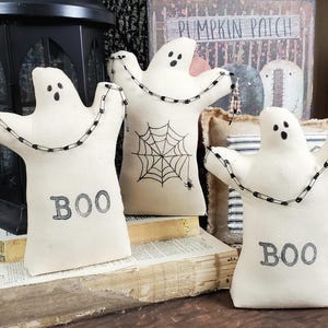 May include: Three white fabric ghost ornaments with black safety pin chains and the word "BOO" embroidered on them. The ghosts are sitting on a stack of old books.