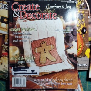 May include: A magazine cover for Create & Decorate, a publication for home decor and crafts. The cover features a gingerbread man applique on a red and white checkered fabric. The text on the cover reads "snuggle into winter... patchwork quilts, felt stockings, wool mittens" and "simple & stylish paper crafts".