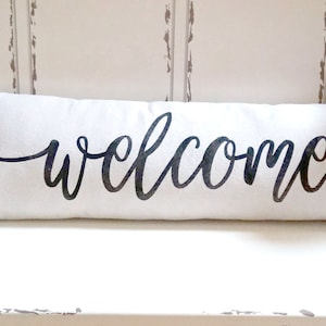 May include: A white lumbar pillow with the word "welcome" written in black cursive script.