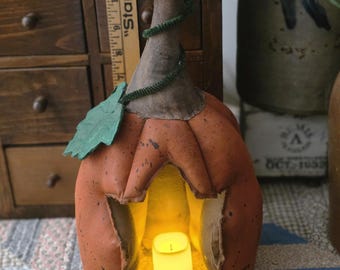 May include: A rustic orange pumpkin-shaped lantern with a cutout face, lit by a warm, glowing candle. The lantern has a brown stem wrapped with green twine and a green leaf. The lantern is sitting on a patterned quilt.