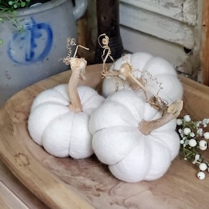 May include: Three white felt pumpkins with brown stems and curly vines sit in a wooden bowl. Small white berries and greenery are arranged nearby. The pumpkins have a ribbed texture, suggesting a fall or harvest theme.