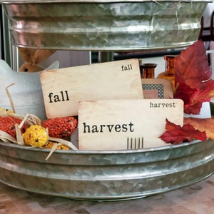 May include: A galvanized metal tiered tray with fall and harvest themed decorations. The tray is filled with faux corn, fall leaves, and two signs that read "fall" and "harvest".
