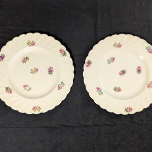 May include: Two white ceramic plates with a scalloped edge and a floral design. The plates have a white background with small pink, purple, and yellow flowers.
