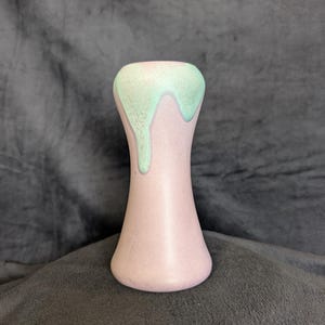 Puede incluir: Un pequeño jarrón de color púrpura claro con una base acampanada y un cuello estrecho. La parte superior del jarrón está decorada con un esmalte verde menta goteante. El jarrón tiene un acabado liso y mate y probablemente sea de cerámica.