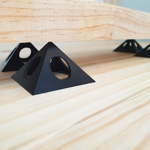 May include: Four black triangular shelf supports with a circular cutout in the center. The supports are made of a hard plastic material and are resting on a light wood surface.