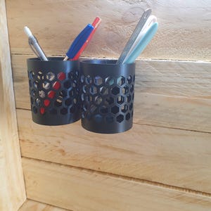 May include: Two black pen holders with a honeycomb design, each holding pens and pencils. The holders are mounted on a light-coloured wooden surface. One holder contains a silver pen, and a red and blue pen. The other holder contains a silver ruler and a light blue pen.