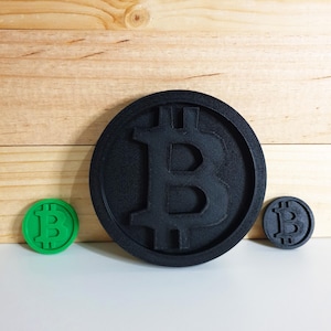May include: Three Bitcoin-themed tokens in black and green. The largest token is black and circular, with a raised Bitcoin symbol. A smaller black token and a green token with the Bitcoin symbol are also present.