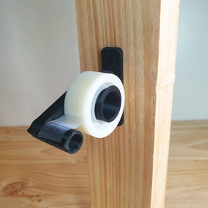 May include: A 3D printed tape dispenser mounted on a wooden surface. The dispenser is black and white and holds a roll of clear tape.