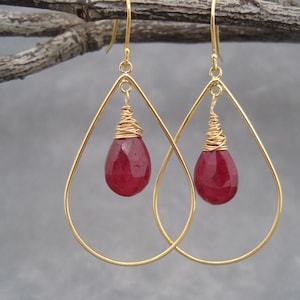 May include: A pair of gold-colored teardrop earrings. Each earring features a faceted, deep red gemstone suspended within a gold wire-wrapped setting. The earrings hang from a thin gold wire.