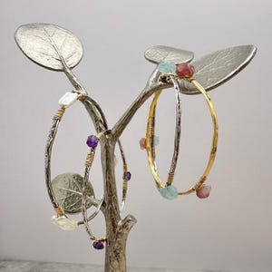 May include: A display of handcrafted bracelets with gemstone accents. The bracelets are silver and gold-toned, adorned with amethyst, pearl, and other colorful stones. They are displayed on a decorative silver tree-shaped stand.