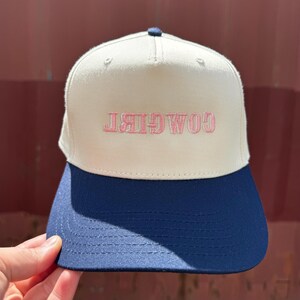 May include: A cream-colored baseball cap with a navy blue brim. The word "COWGIRL" is embroidered in pink across the front. The cap has a structured fit and a curved brim.