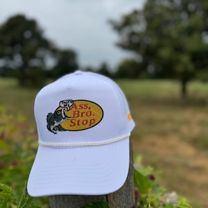Puede incluir: Gorra de camionero blanca con borde de cuerda y un gráfico en la parte delantera. El gráfico es un óvalo con un pez y el texto "That's My Ass, Bro. Stop". La gorra tiene una corona blanca y una visera curva.