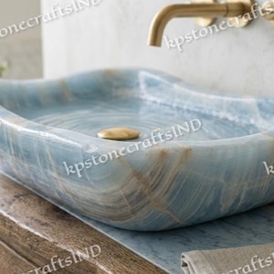 May include: A rectangular vessel sink crafted from light blue stone with natural white and tan veining. The basin features smooth, rounded edges and a polished finish, complemented by a centered gold-toned drain cover and a wall-mounted gold faucet.