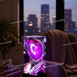 May include: A rectangular, illuminated lamp with a striking purple agate design. The lamp sits on a black base and is placed on a dark wooden table. The background features a city skyline at dusk, with a cozy chair and blanket.