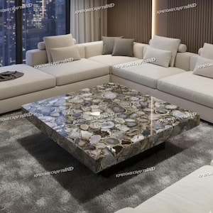 May include: A square coffee table with a polished stone top featuring a mosaic of agate slices in shades of gray, brown, and white. The table sits on a black base, and is surrounded by a cream-colored sectional sofa.