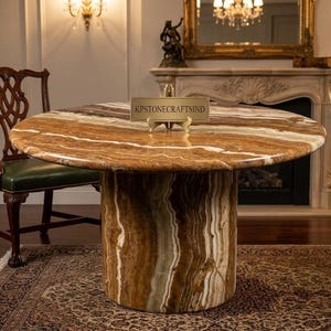 May include: A round, brown and tan marble table with a cylindrical base. The table has a polished surface and is set in a room with a fireplace and a decorative chair. The table has a sign that reads "KPSTONECRAFTSIND".