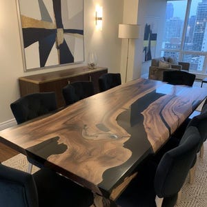 Op de afbeelding: Een grote, rechthoekige eettafel met een houten blad met levende rand en zwarte harsinleg. Acht zwarte fluwelen stoelen omringen de tafel. De kamer heeft een modern abstract schilderij en uitzicht op de stad.