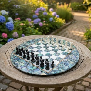 Puede incluir: Un tablero de ajedrez redondo con borde de nácar sobre una mesa de madera. El tablero tiene un patrón de cuadros blancos y negros. Las piezas de ajedrez blancas y negras están dispuestas. El fondo muestra un jardín con flores coloridas.