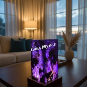 May include: A rectangular purple lamp with the text "Casa Mytics" on the front. The lamp sits on a dark brown wooden base and is on a dark brown table. The lamp is in a well-lit room with a couch and windows.