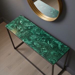 Puede incluir: Una consola rectangular con una tapa verde malaquita y un marco de metal oscuro. Un espejo redondo con marco dorado cuelga de la pared gris sobre la mesa. La mesa está colocada sobre un suelo de madera clara.