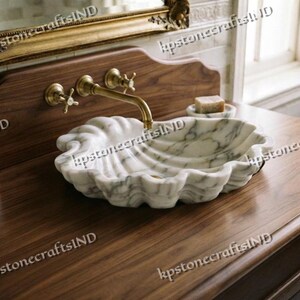 May include: A white marble vessel sink shaped like a large seashell, featuring grey veining and a scalloped edge. The sink is mounted on a dark brown wooden vanity countertop with a brass wall-mounted faucet and two matching cross-handle knobs.