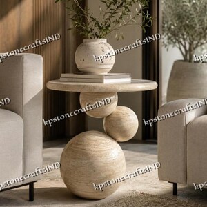 May include: A round beige travertine side table featuring a stacked design with three spherical bases of varying sizes supporting a flat circular tabletop. A matching beige stone vase with green foliage sits on top of the table.