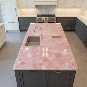 Puede incluir: Isla de cocina con encimera rosa y fregadero de acero inoxidable. La encimera tiene un aspecto veteado con tonos blancos y rosas. La base de la isla es gris oscuro con cajones y tiradores. Se instala un grifo de acero inoxidable.