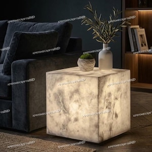 May include: A square, illuminated side table with a marble-like appearance. The table is a cube shape and has a soft, glowing light. A small potted plant and a white vase with greenery sit on top. The text "KPSTONECRAFTSIND" is visible on the table.