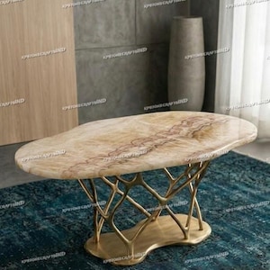 Op de afbeelding: Een ovale salontafel met een lichtbruin marmeren blad en een goudkleurige, takachtige basis. De tafel staat op een blauwgroen tapijt. Het marmeren blad heeft roodbruine aderen. De basis is gemaakt van verweven metaal.