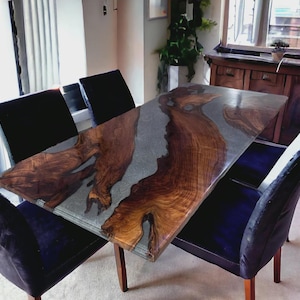 Puede incluir: Una mesa de comedor rectangular con un diseño de borde vivo de madera y un centro de resina epoxi gris. La mesa está rodeada por cuatro sillas tapizadas de color azul oscuro. La mesa mide aproximadamente 183 cm de largo.