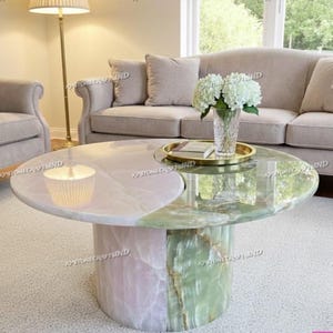 May include: A unique, two-tone coffee table with a round top. One half is a light pink, the other is green. The base is a matching pink and green. The table is in a living room with a sofa and chair.