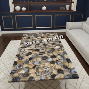 Puede incluir: Una mesa de centro rectangular con una tapa de piedra de mosaico en tonos beige, gris y negro. La mesa tiene una base transparente y moderna y está situada en un salón con una alfombra y un sofá. El texto "KPSTONECRAFTSIND" es visible.