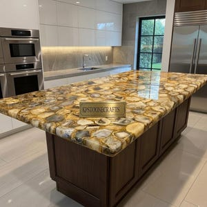 May include: A large kitchen island with a countertop made of polished, irregularly shaped, golden-brown and gray stones. The island has a dark brown wooden base and a brass sign that reads "KPSTOONECRAFTS". The kitchen features white cabinets and stainless steel appliances.