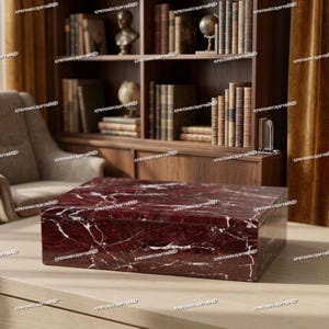 Può includere: Una scatola rettangolare in marmo bordeaux con venature bianche è appoggiata su una superficie chiara. La scatola è davanti a una libreria in legno piena di libri e oggetti decorativi. La scatola di marmo è un contenitore.