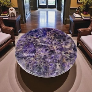 May include: A round coffee table with a polished amethyst top. The table's surface displays a mix of purple, white, and gold hues. The table is set in a living room with two chairs and side tables.
