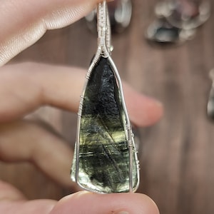 May include: A dark green, teardrop-shaped pendant with a silver wire wrap. The stone has a textured surface and is held by a person's fingers. Other pendants are visible in the background.