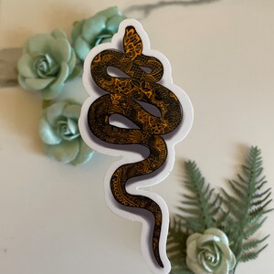 Op de afbeelding: Een zwarte en gouden slangen sticker met ingewikkelde details. De slang is opgerold in een spiraal met zijn kop omhoog. De sticker is op een witte achtergrond.