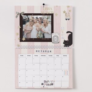 May include: A pink and white striped wall calendar for October, featuring a photo, candle, and cat illustration. The calendar includes the text "Libra Season" and "Happy B'day". The calendar is spiral-bound.