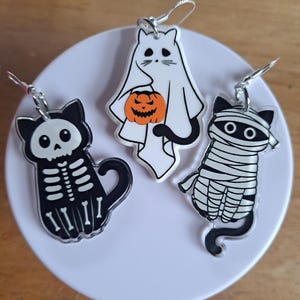 May include: Three Halloween-themed earrings. One is a black cat skeleton, another is a white ghost holding an orange jack-o'-lantern, and the third is a mummy cat. All have silver-colored ear wires.