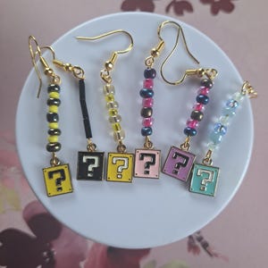 May include: A collection of handmade earrings with gold-colored hooks. Each earring features a square charm with a question mark and colorful beaded accents. The charms are in yellow, black, pink, purple, and teal.